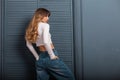 Fashionable slender young woman with long red hair in a white fashionable top in stylish denim pants poses near a vintage blue