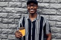 Fashionable smiling african american man in hat, man outdoors with takeaway coffee cup, over gray wall background Royalty Free Stock Photo