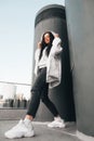 Fashionable street portrait of a girl in casual clothing, wearing a white jacket, sneakers and glasses, stands in full height,