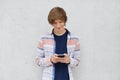 Fashionable teenage boy wearing shirt, holding cell phone in hands, messaging with friends or playing games online using free inte