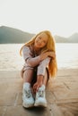 Fashionable teenage girl in a long dress and sneakers. Cute long-haired girl resting on the beach near mountains and ocean Royalty Free Stock Photo