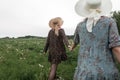 Fashionable woman in black stylish dress in fashion straw vintage hat holds girlfriends hand along field with green grass. Two Royalty Free Stock Photo