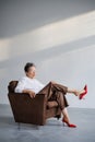 Fashionable mature woman in red heels and white shirt sitting on armchair on grey background Royalty Free Stock Photo