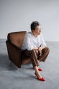 Fashionable mature woman in red heels and white shirt sitting on armchair on grey Royalty Free Stock Photo