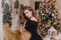 Fashionable woman with wineglass sitting on brown sofa and posing near christmas tree. Indoor photo of graceful lady in