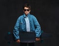 Fashionable young guy in sunglasses dressed in a t-shirt and denim jacket holds a longboard. Royalty Free Stock Photo