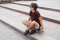 Fashionable young hipster man in black sunglasses in a vintage t-shirt in trendy shorts in sneakers resting while sitting Royalty Free Stock Photo