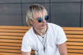 Fashionable young hipster man in blue sunglasses in white t-shirt sitting on a wooden bench near a gray building on a summer day. Royalty Free Stock Photo