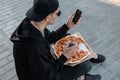 Fashionable young hipster man in stylish black clothes with a pizza with a glass bottle sits on the sidewalk