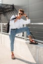 Fashionable young hipster man in stylish white and denim clothes in red leather sandals in sunglasses with a fabric bag rest Royalty Free Stock Photo