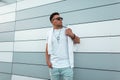 Fashionable young hipster man with trendy hairstyle in sunglasses in jeans in a white T-shirt posing near modern building on the Royalty Free Stock Photo