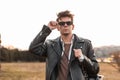 Fashionable young hipster man in vintage leather oversized black jacket with backpack straightens sunglasses outdoors. American Royalty Free Stock Photo