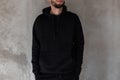 Fashionable young man with a beard in black stylish clothes is standing indoors near a concrete vintage wall. Trendy cool guy. Royalty Free Stock Photo