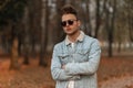 Fashionable young man with a beard with a stylish hairstyle in trendy sunglasses in a blue denim jacket is relaxing outdoors in Royalty Free Stock Photo