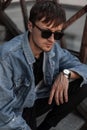 Fashionable young man hipster in stylish denim clothes trendy in a black sunglasses sits on a vintage staircase in the city Royalty Free Stock Photo