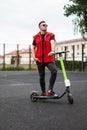 Fashionable young man in a red vest in black jeans in sneakers in sunglasses stands with a modern electric scooter Royalty Free Stock Photo