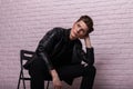 Fashionable young man in a trendy black leather jacket is sitting on a chair near a pink vintage brick wall indoors. Stylish Royalty Free Stock Photo