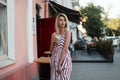 Fashionable young positive woman in a stylish pink striped sundress stands with a tasty milkshake in hands. Cheerful girl