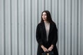 Fashionable young woman with brown hair in black fashionable coat with stylish leather black handbag stands near silver metal