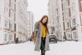 Fashionable young woman in coat with backpack walking on street in big city in snowing time. Cheerful mood, snowfall