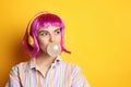 Fashionable young woman in colorful wig with headphones blowing bubblegum on yellow background, space for text Royalty Free Stock Photo