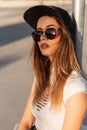 Fashionable young woman hipster in fashion casual t-shirt in trendy baseball black cap in stylish sunglasses sit on asphalt at Royalty Free Stock Photo