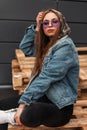 Fashionable young woman in purple glasses in stylish clothes puts military hood on head. American trendy hipster girl in youth Royalty Free Stock Photo