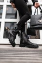 Fashionable young woman stands on the stone steps in the city in black trendy leather boots in vintage jeans with a bag. Close up