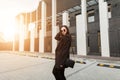 Fashionable young woman in stylish black blazer with vintage handbag walks near building in city at sunset. Urban girl travels and Royalty Free Stock Photo