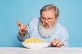 Fastidious senior hairy gray-bearded man tasting large portion of noodles, pasta isolated on blue studio background.