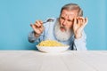 Fastidious senior hairy gray-bearded man tasting large portion of noodles, pasta on blue studio background.