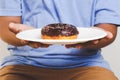 Fat Asian kid is holding a white plate with chocolate glazed donut on it. Royalty Free Stock Photo