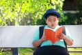 Fat boy wearing a blue shirt wearing a hat Open bench reading book Bench in garden under tree.