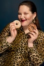 A fat girl in a leopard blouse holds donuts near her face. Dark blue background.