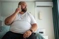 Fat guy calls on phone, sitting on bed, looking out window, signs up for gym to put figure in order Royalty Free Stock Photo