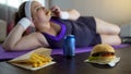 Fat lazy girl in sportswear eating junk food instead of training on yoga mat