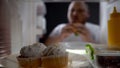 Fat man eating burger in front of fridge, junk food addiction, cupcakes closeup Royalty Free Stock Photo