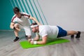 Fat man looks exhausted while doing exercises standing in bar on the mat in the fitness gym under strict control of Royalty Free Stock Photo