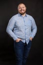 A fat red-haired man with a beard and mustache in a blue shirt and jeans stands holding his hands in his pockets and laughs. Black Royalty Free Stock Photo