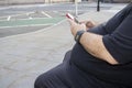 A fat unhealthy overweight man sitting on a park bench texting on his phone