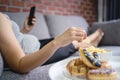 Fat woman lying on sofa holding smartphone and eating potato chips, overeating concept