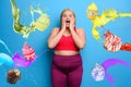 Fat woman with shocked and surprised expression because she is surrounded by sweets. Cyan background