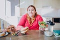 Fat young woman in kitchen sitting and eating food. Confused unhappy plus size model hold burger and bowl salad in hands