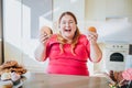 Fat young woman in kitchen sitting and eating junk food. Happy excited plus size model laughing. Holding burgers in Royalty Free Stock Photo