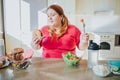Fat young woman in kitchen sitting and eating junk food. Holding burger and tomato on fork. Regret and unhappiness. Body