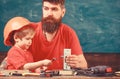 Father with beard teaching little son to use tools in classroom, chalkboard on background. Mens work concept. Boy, child