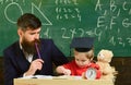 Father checking hometask, helps to boy, son. Teacher in formal wear and pupil in mortarboard in classroom, chalkboard on Royalty Free Stock Photo