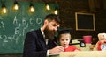 Father checking homework, helps to boy, son. Teacher in formal wear and pupil in mortarboard in classroom, chalkboard on Royalty Free Stock Photo