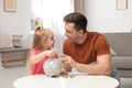 Father and daughter putting coin into piggy bank at table indoors. Royalty Free Stock Photo