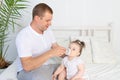 A father feeds his little daughter from a bottle on a white bed at home, the concept of baby food and child care